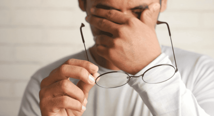 Stressful man holding glasses and covering face in frustration A man in a white shirt holding glasses and covering his face with his hand, showing frustration.